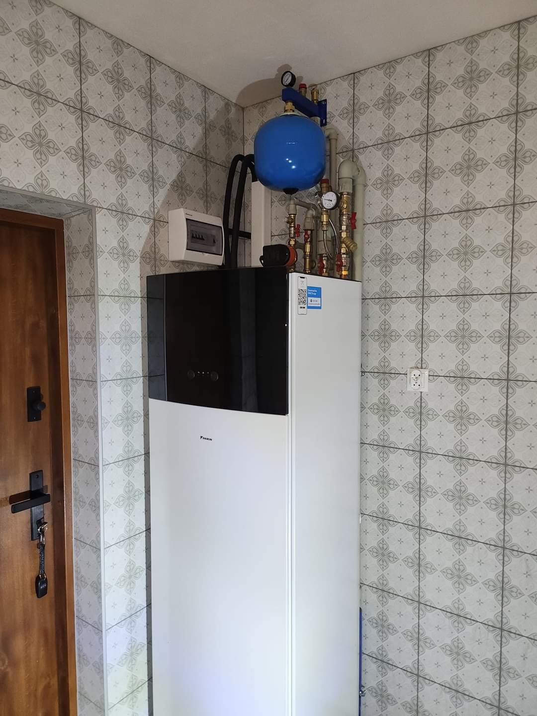 Biała pompa ciepła Daikin z czarnym panelem górnym, niebieski zbiornik wyrównawczy i instalacja hydrauliczną na tle ściany w płytkach z wzorem w kształcie kwiatów.