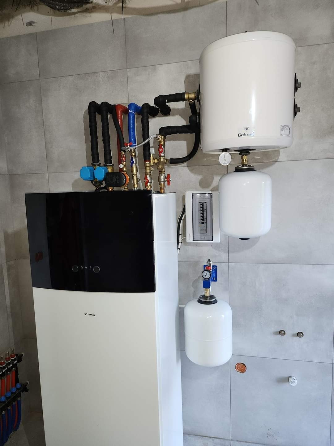 Biała pompa ciepła Daikin z czarnym panelem górnym, podłączona do białego zasobnika Galmet i dwóch zbiorników wyrównawczych, na tle szarych płytek ceramicznych.