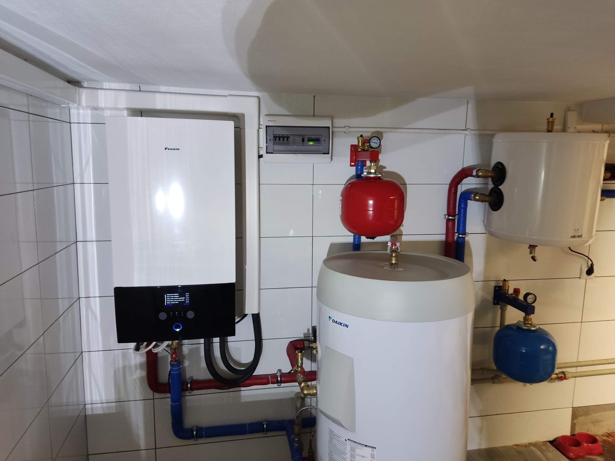 Instalacja pompy ciepła Daikin z zbiornikiem buforowym, rurami w kolorze czerwonym i niebieskim, oraz dodatkowym zbiornikiem wyrównawczym w pomieszczeniu wyłożonym białymi płytkami.