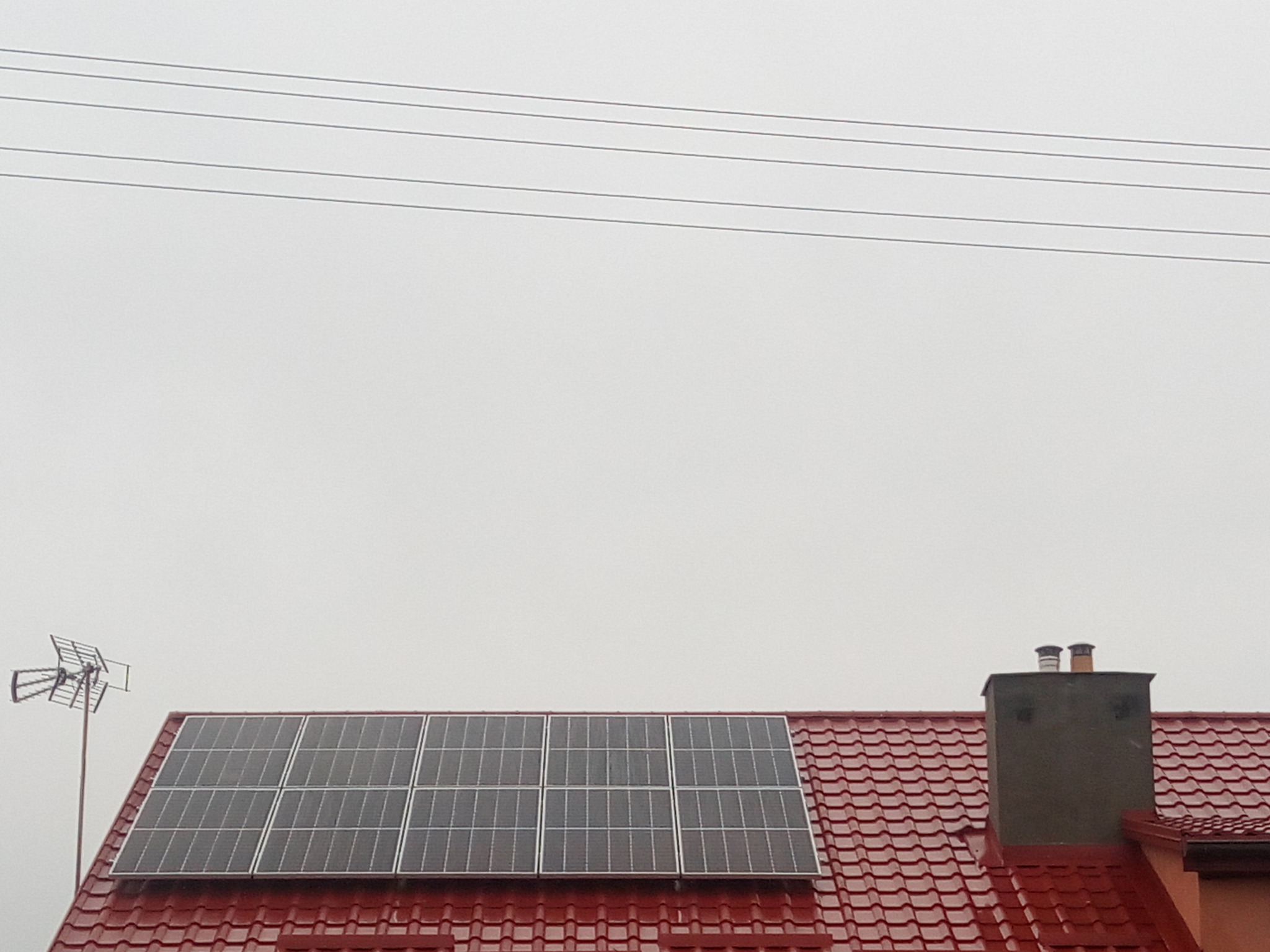 Panele słoneczne na czerwonym dachu domu jednorodzinnego, obok komin i antena telewizyjna, pochmurne niebo w tle.