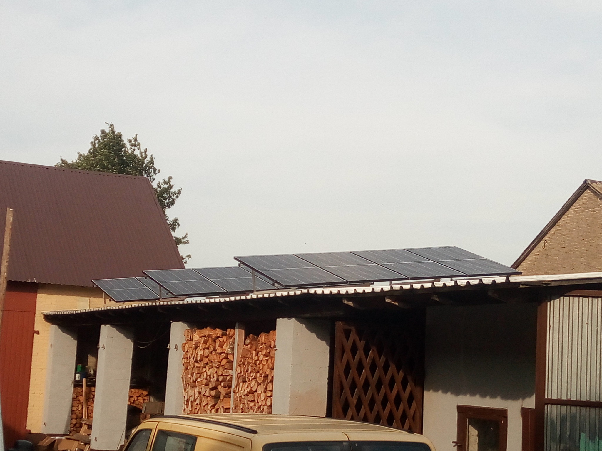 Panele słoneczne zamontowane na dachu budynku gospodarczego z zapasem drewna, widoczne zza zaparkowanego samochodu.