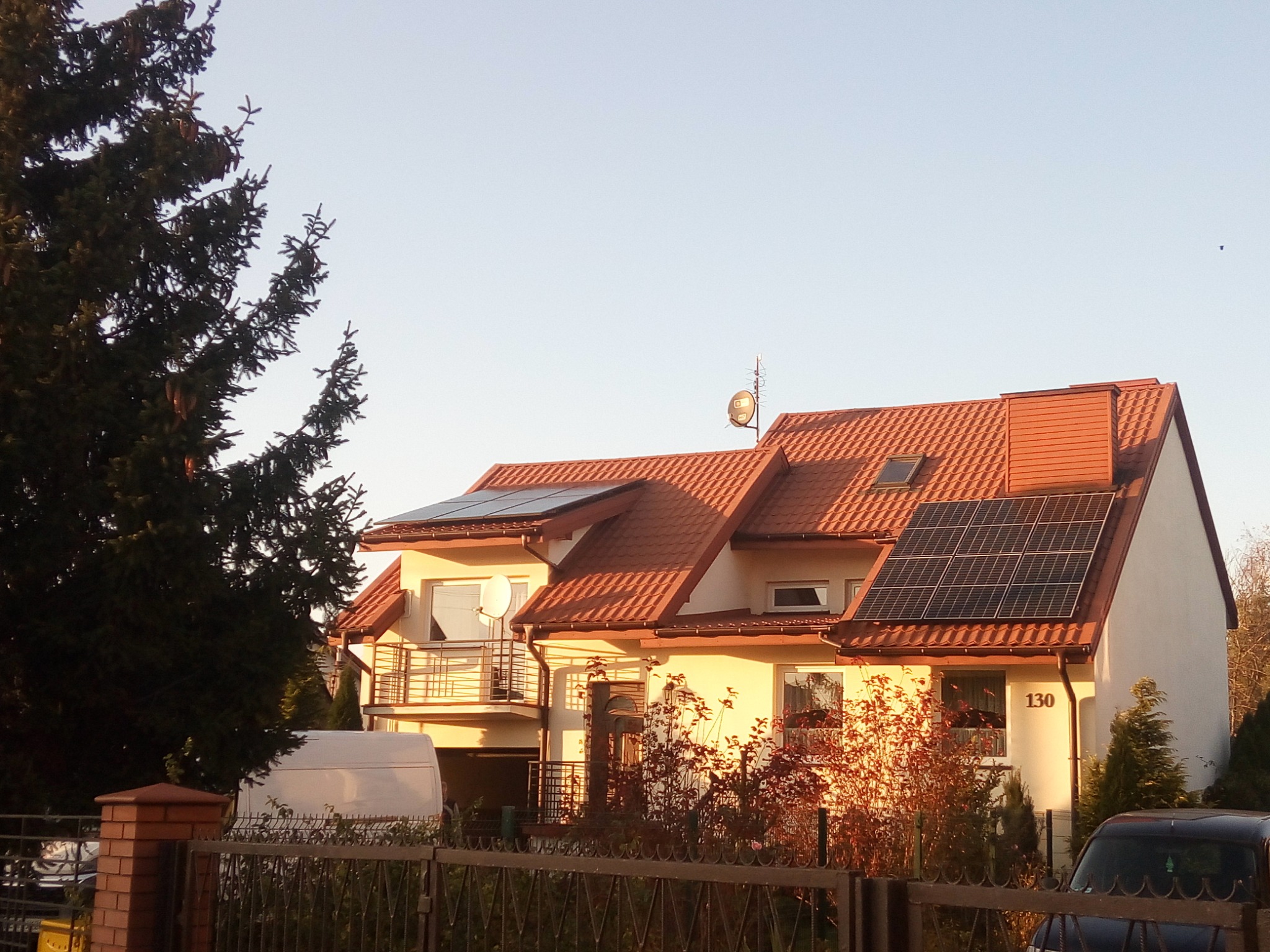 Dwukondygnacyjny dom jednorodzinny z brązowym dachem i panelami fotowoltaicznymi na dachu, widoczny zza ogrodzenia z bliska, w tle zaparkowany biały bus i czarny samochód.