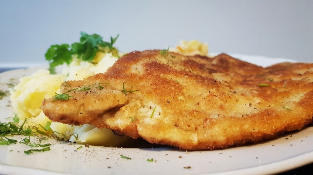 Panierowany kotlet schabowy z ziemniaczanym puree i zieloną pietruszką na białym talerzu, delikatnie posypany koperkiem.