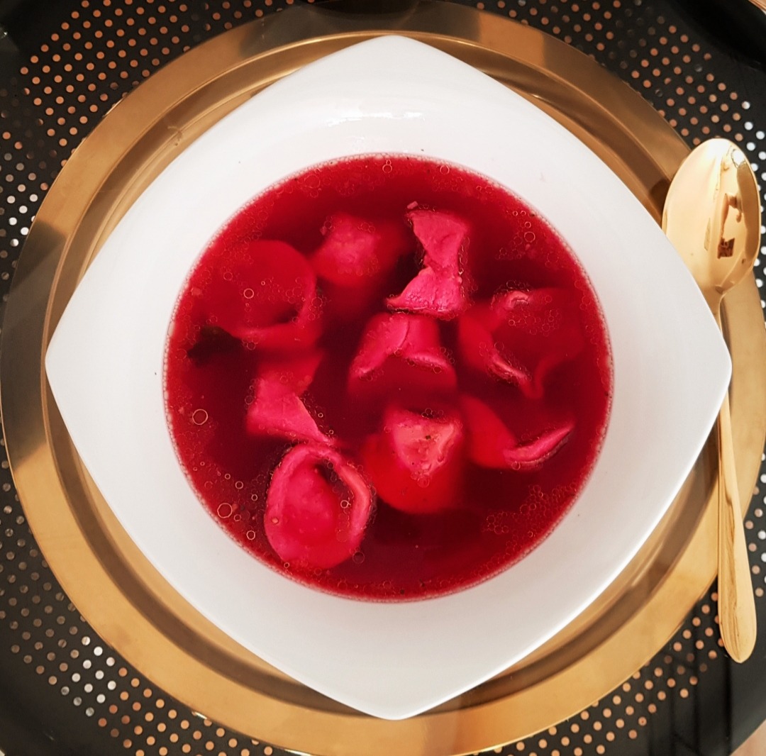 Bursztynowy bulion z uszkami w białym kwadratowym talerzu na złotej tacy, złota łyżka obok.