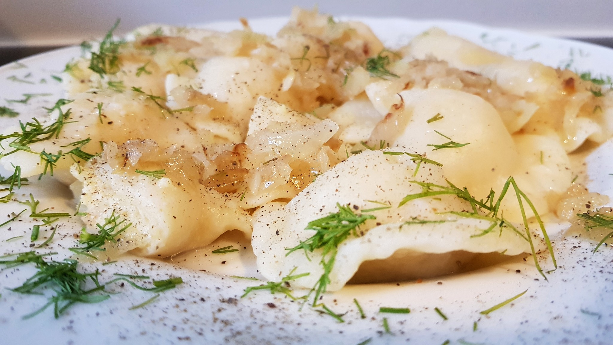 Porcja pierogów z kapustą i grzybami, polanych podsmażoną cebulką i posypanych świeżym koperkiem, podane na białym talerzu.