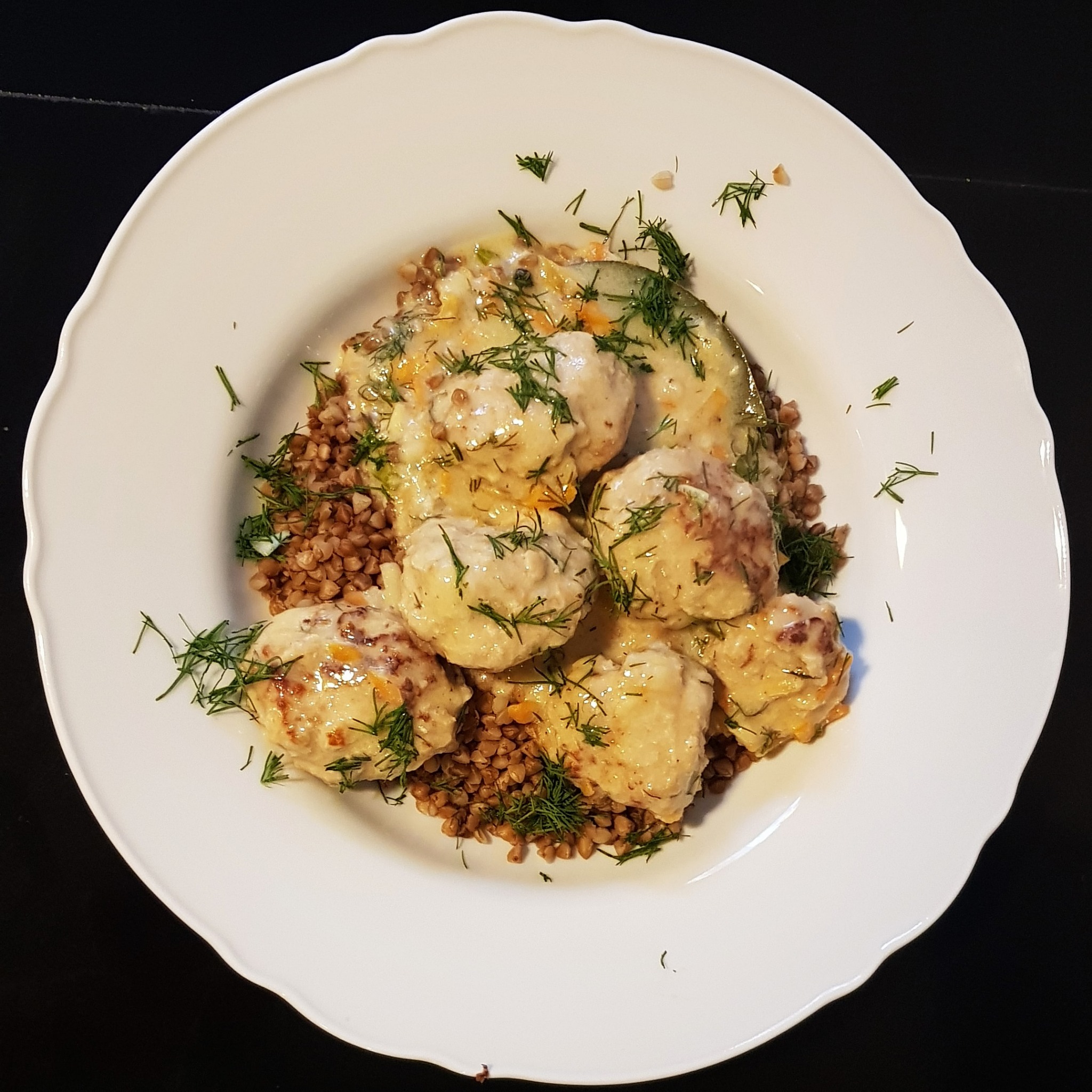Porcja pulpetów w kremowym sosie, podana na kaszy gryczanej, udekorowana świeżym koperkiem na białym talerzu z falistym brzegiem, na czarnym tle.