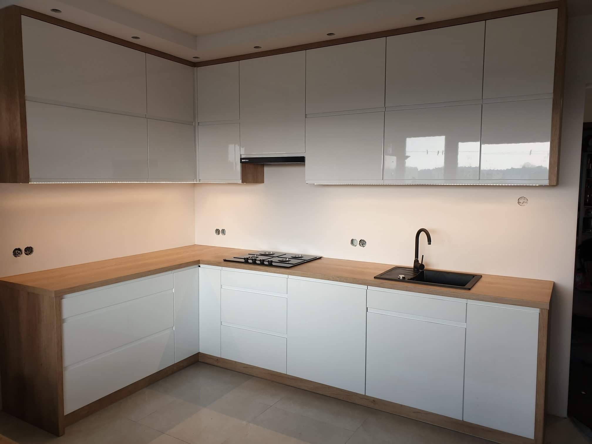 Nowoczesna kuchnia z białymi, lakierowanymi frontami i drewnianym blatem, czarna armatura, płyta grzewcza, oświetlenie LED pod szafkami wiszącymi, widoczne niedokończone gniazdka elektryczne.