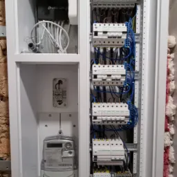 Wewnętrzny widok rozdzielnicy elektrycznej w białej obudowie, z licznikiem energii, zabezpieczeniami nadprądowymi i modułem TP-Link, zamontowanej na ścianie z izolacją.