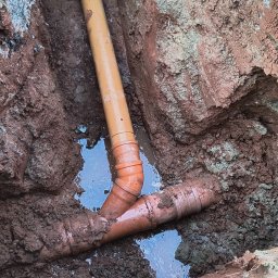 P.B. Marcin Chrzanowski - Pomarańczowe rury kanalizacyjne ułożone w wykopie, częściowo zanurzone w wodzie, z widocznymi śladami błota i ziemi.