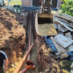 P.B. Marcin Chrzanowski - Wykopany rów pod instalację z ułożonymi pomarańczowymi rurami kanalizacyjnymi, widoczna łyżka koparki z ziemią i stos desek w tle, budynek w trakcie budowy.