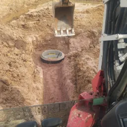 Głęboki wykop w ziemi z betonowym kręgiem studziennym w środku, widok z kabiny koparki, z widocznym łyżką koparki nad wykopem i elementami wnętrza kabiny na pierwszym planie.
