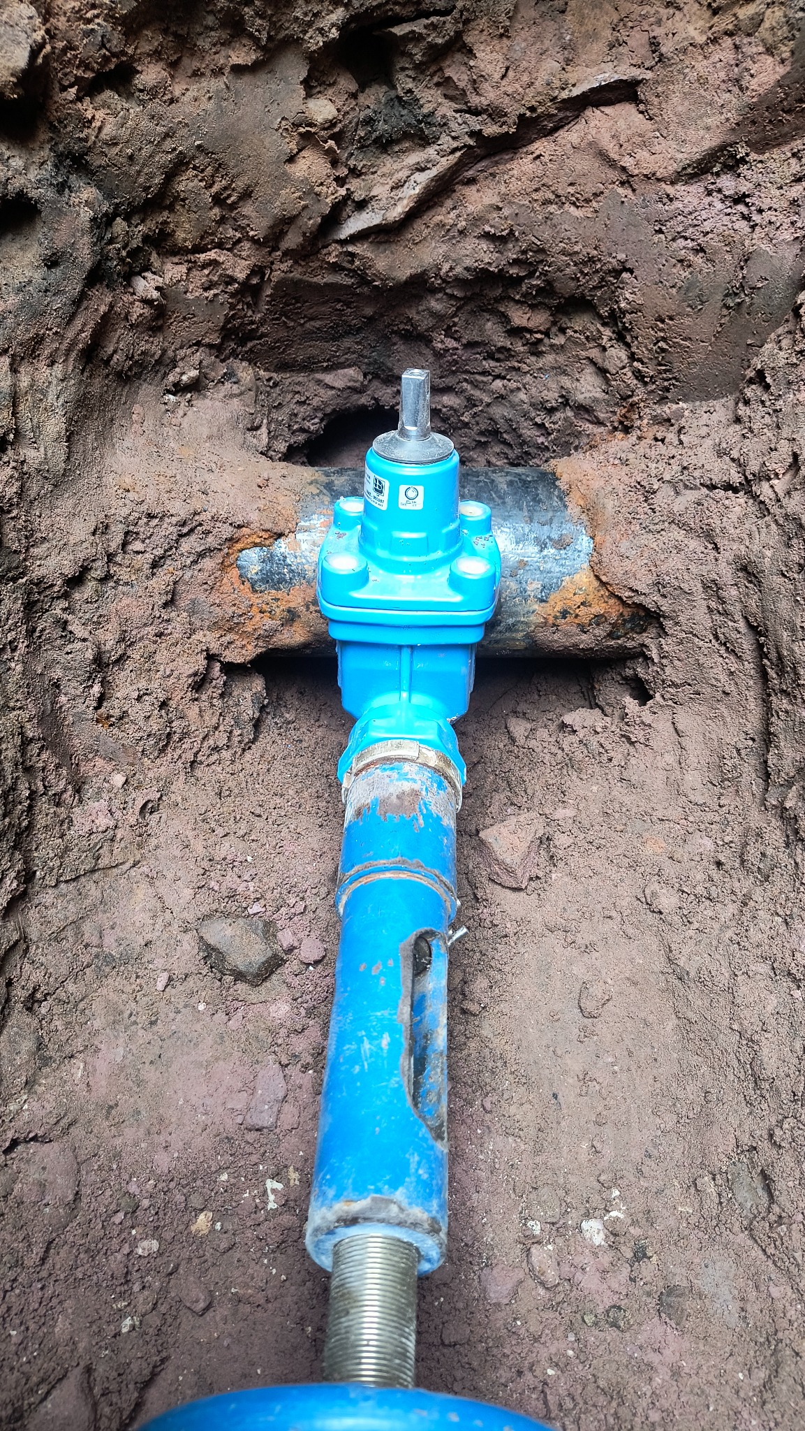 Niebieski zawór przyłącza wodno-kanalizacyjnego w wykopie ziemnym w Zagnańsku, widoczne gwintowane zakończenie rury i fragment skorodowanej rury głównej.
