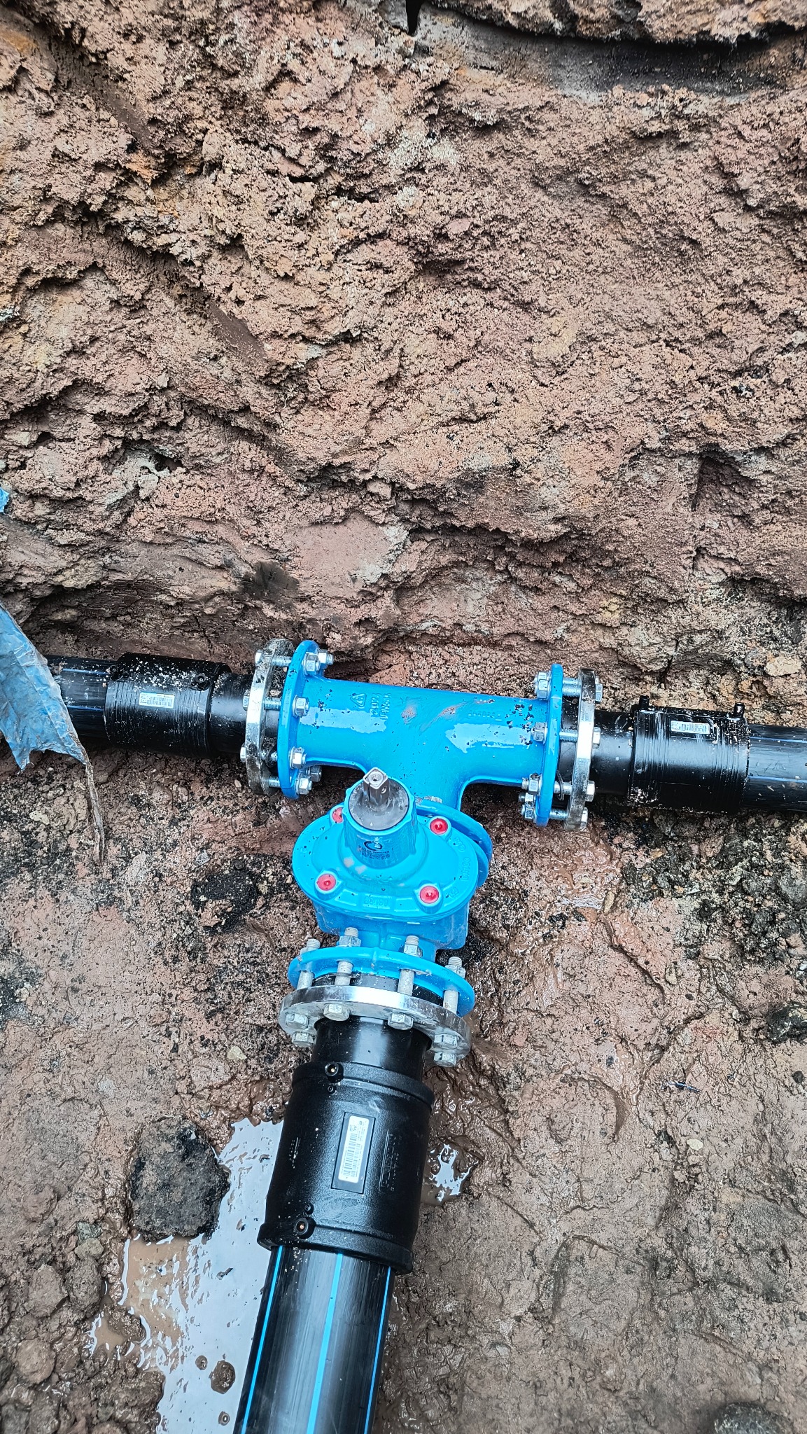 Niebieski zawór trójnikowy z kołnierzami, połączony z czarnymi rurami w wykopie z ziemi gliniastej, widoczne ślady wilgoci.