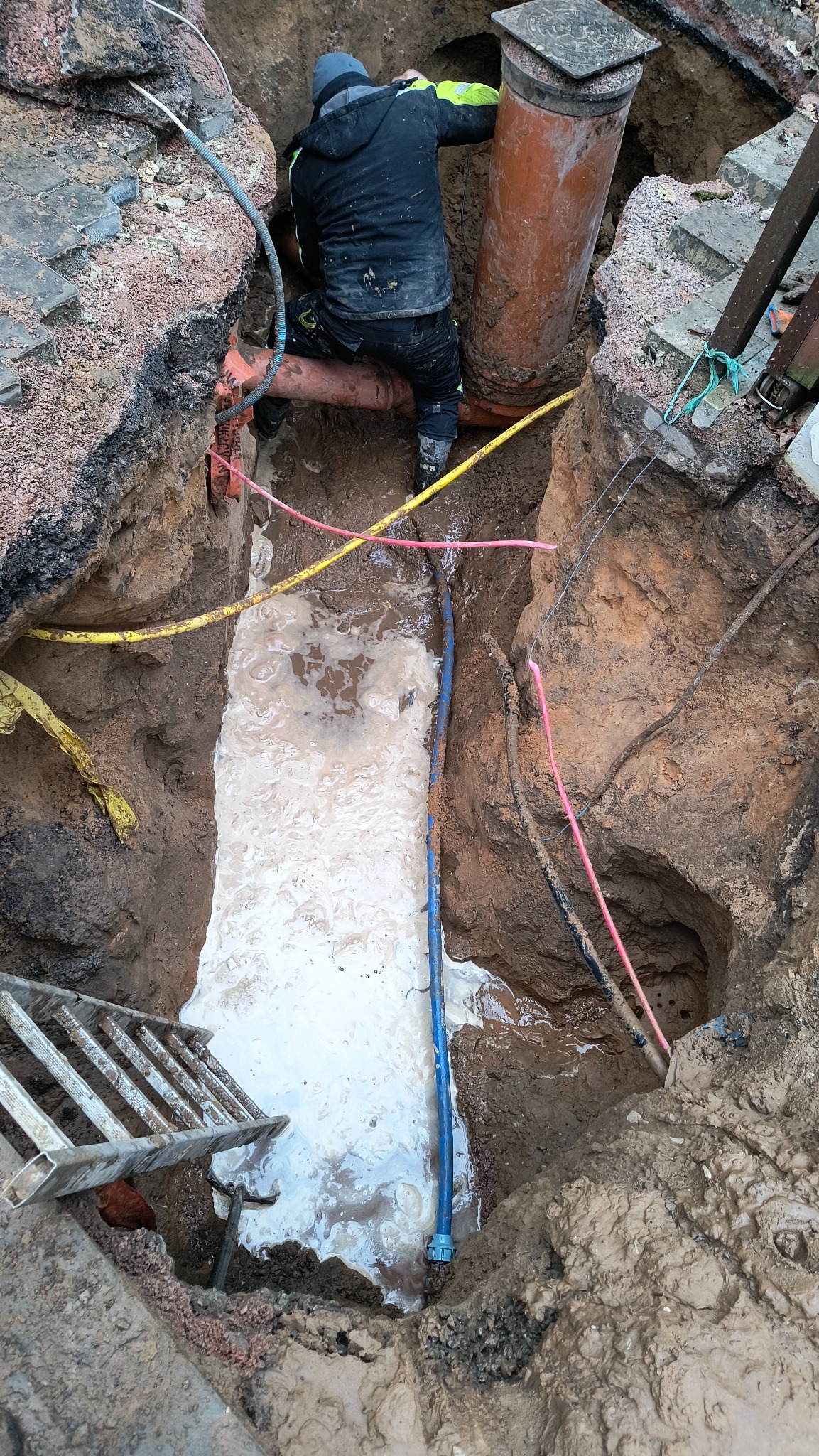 Pracownik w wykopie podczas montażu rur, widoczne kable, błoto i drabina, duża rura kanalizacyjna obok