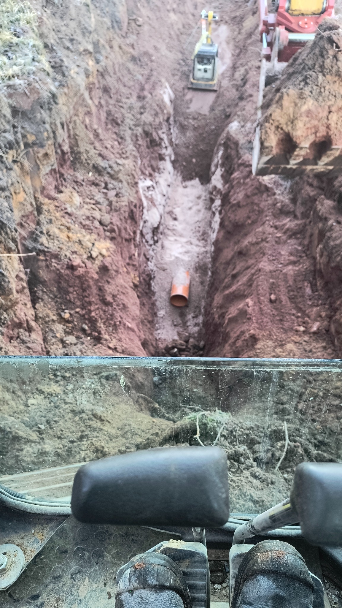 Wykopany rów w ziemi z pomarańczową rurą na dnie, widok z kabiny koparki z widocznymi butami operatora i elementami sterującymi, w tle pracują maszyny budowlane.