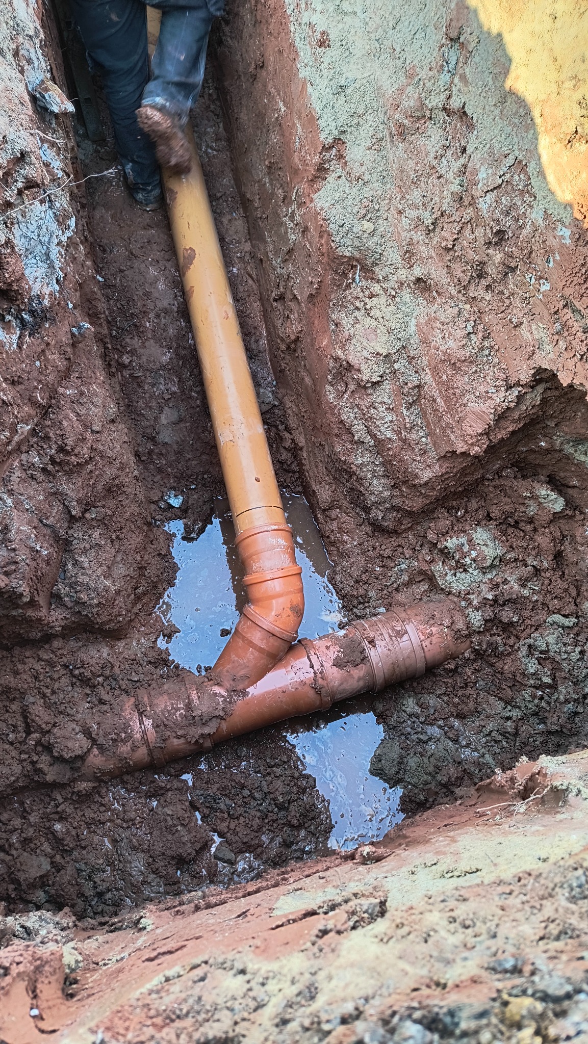 Pomarańczowe rury kanalizacyjne ułożone w wykopie, częściowo zanurzone w wodzie, z widocznymi śladami błota i ziemi.