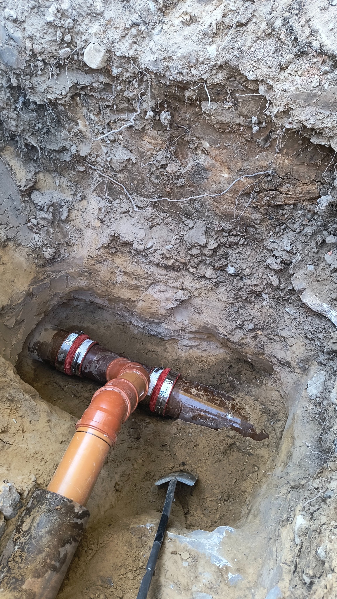 Połączenie rur kanalizacyjnych w wykopie ziemnym, widoczne obejmy zaciskowe i szpadel w tle.