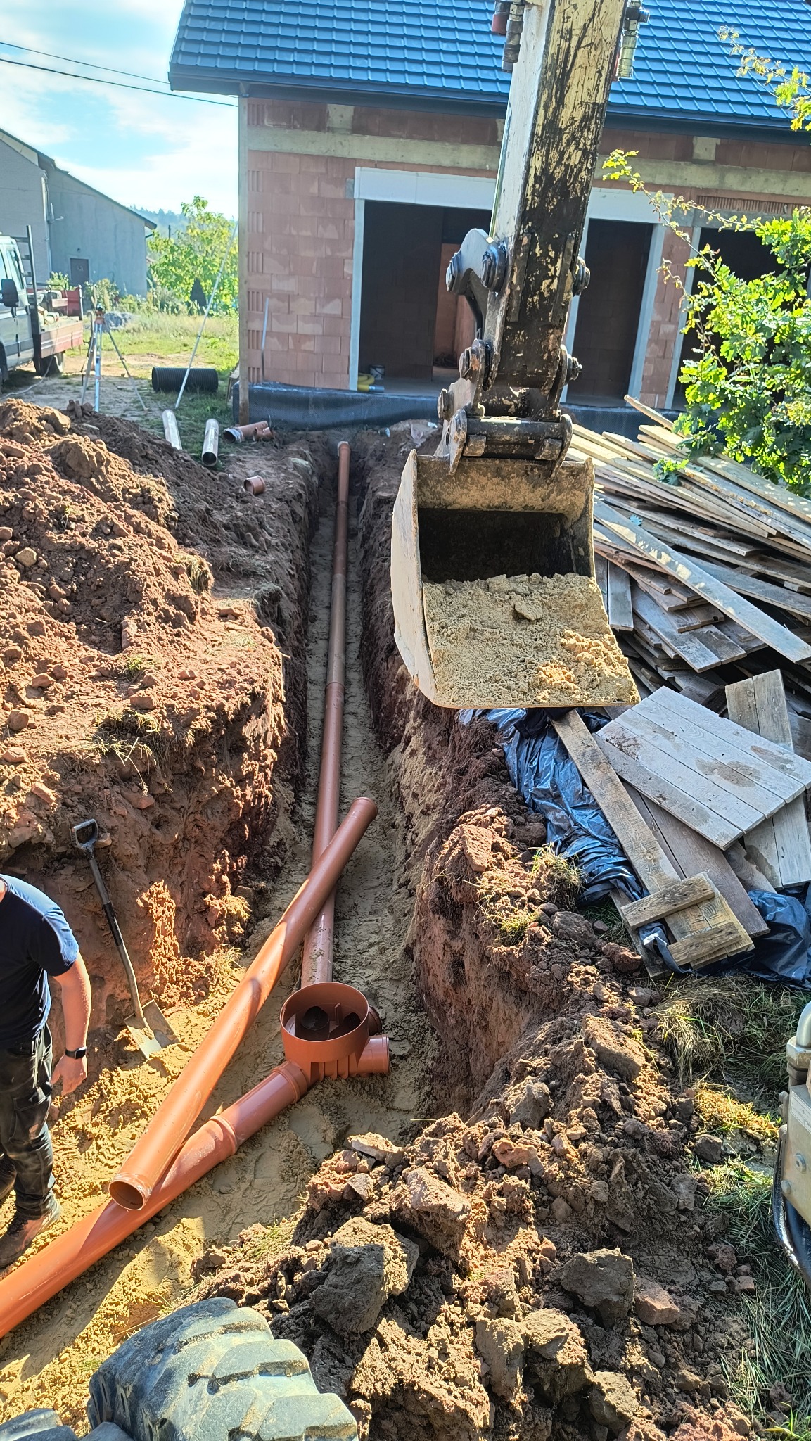 Wykopany rów pod instalację z ułożonymi pomarańczowymi rurami kanalizacyjnymi, widoczna łyżka koparki z ziemią i stos desek w tle, budynek w trakcie budowy.