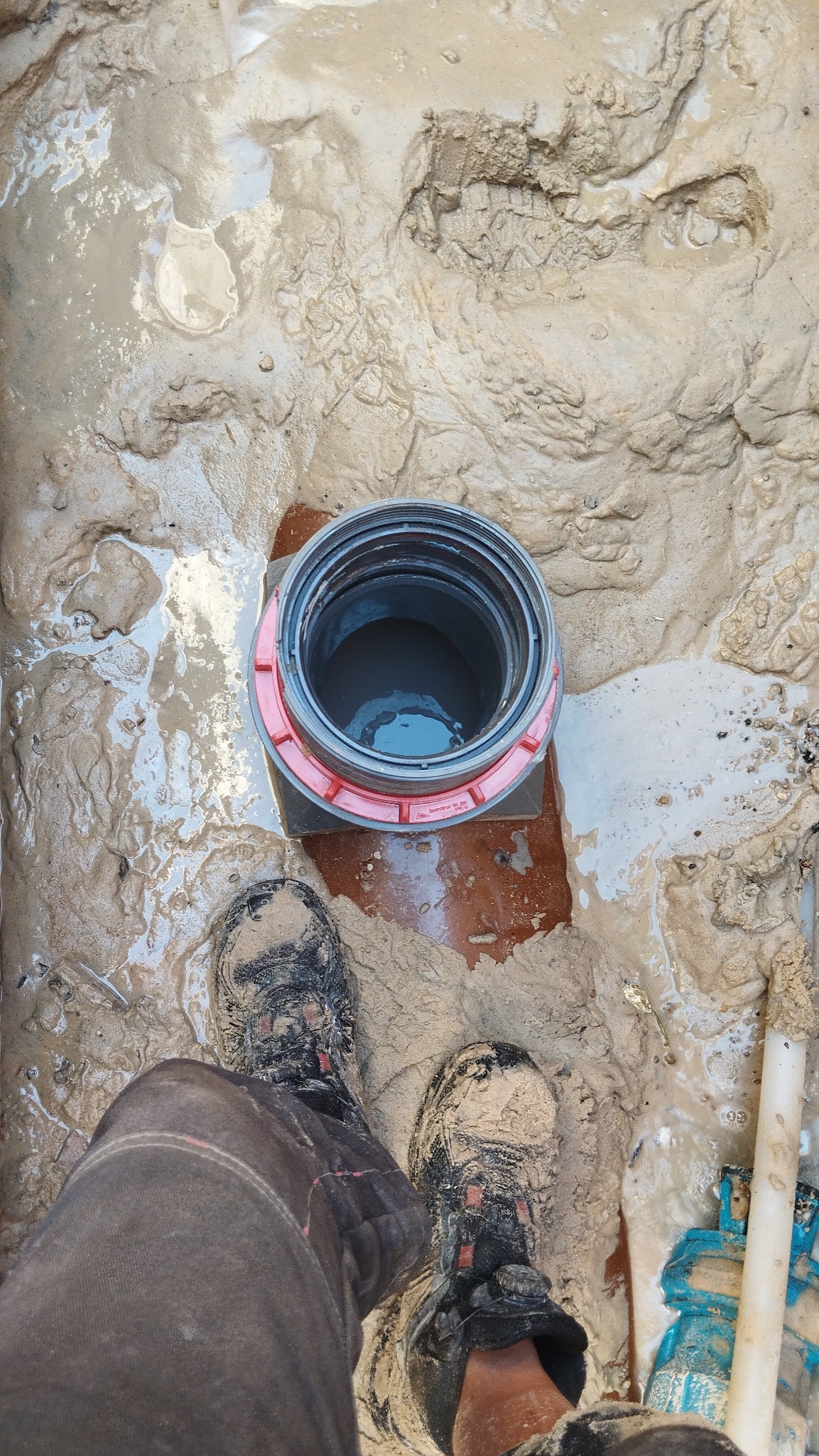 Otwór kanalizacyjny z widocznymi śladami butów w błotnistym wykopie, fragment rury i narzędzia w tle.
