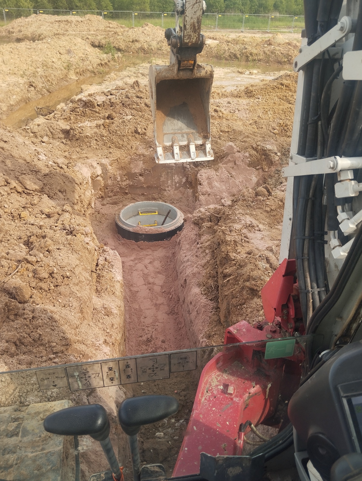 Głęboki wykop w ziemi z betonowym kręgiem studziennym w środku, widok z kabiny koparki, z widocznym łyżką koparki nad wykopem i elementami wnętrza kabiny na pierwszym planie.