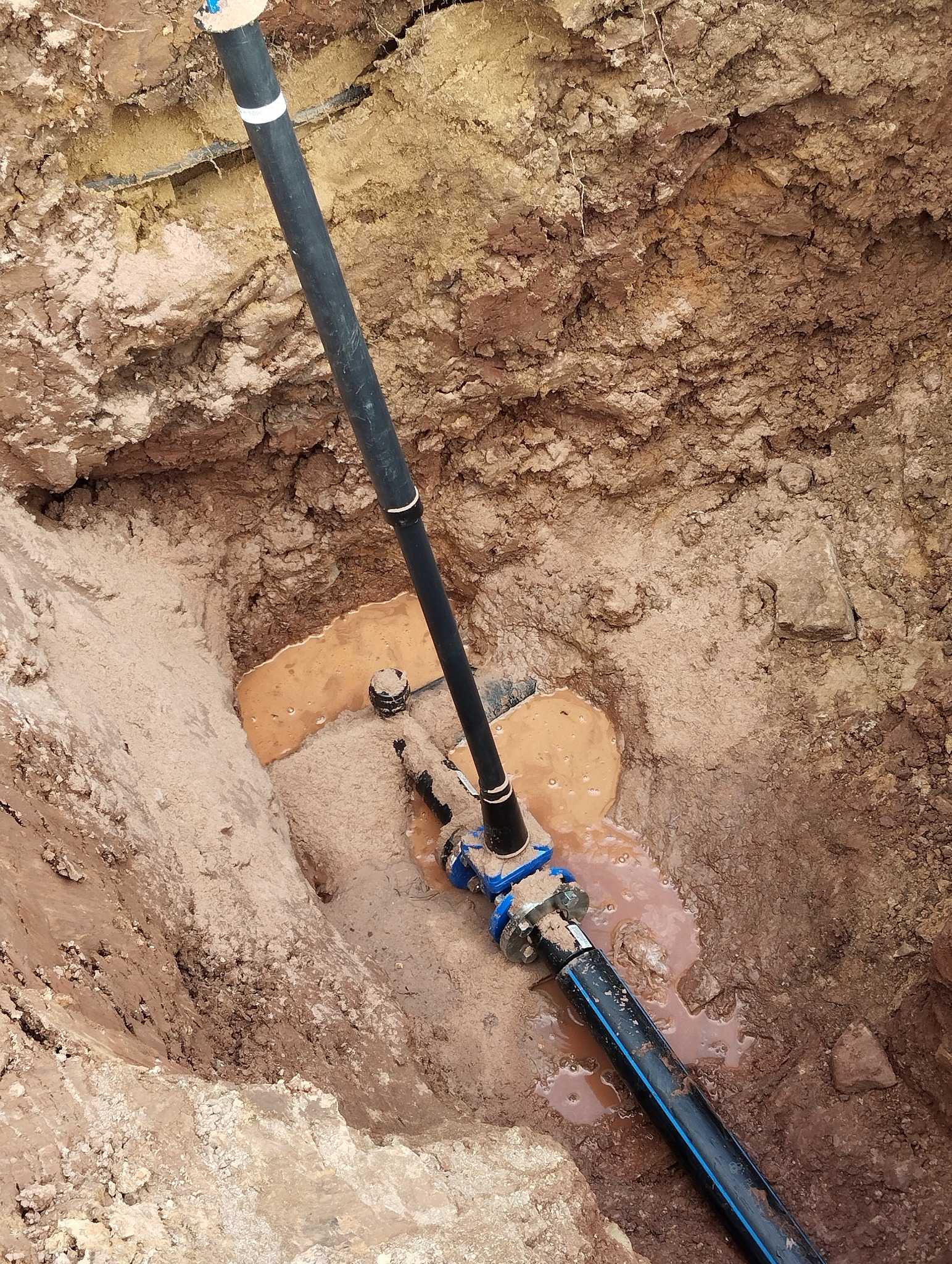 Połączenie rur wodociągowych z niebieskimi złączkami w wykopie wypełnionym wodą, widoczne warstwy ziemi.