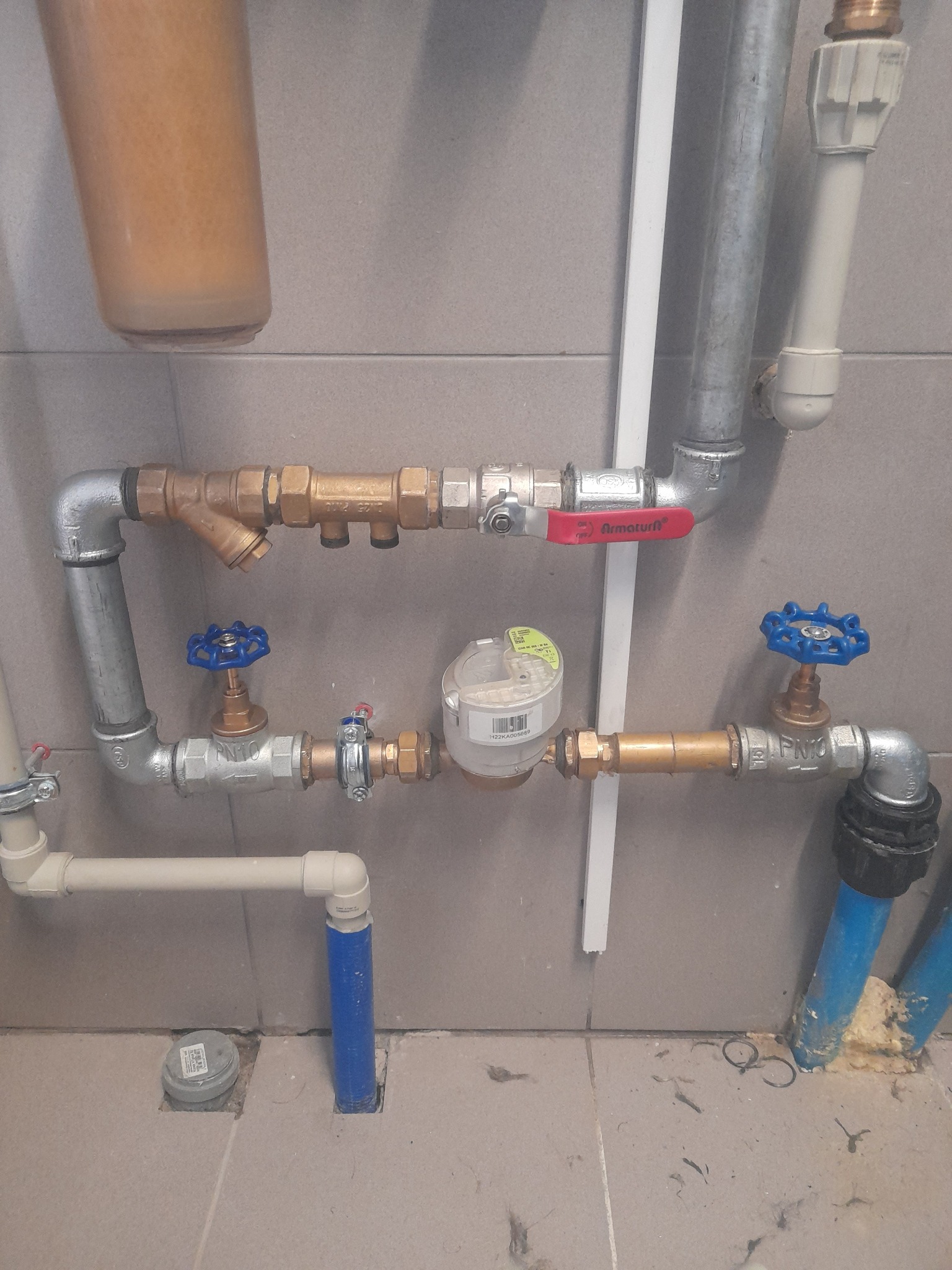Kompleksowy system rur wodociągowych z filtrem, zaworami z niebieskimi pokrętłami i licznikiem zużycia wody, zainstalowany wewnątrz budynku na tle płytek ceramicznych.