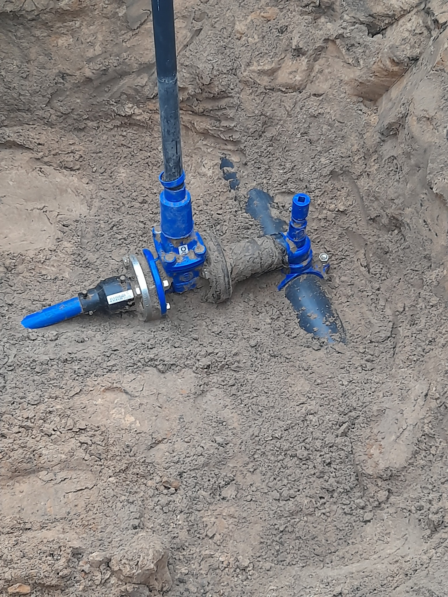 Niebieskie zawory i rury w wykopie ziemnym, widoczne połączenia i elementy instalacji, ziemia pokrywa częściowo elementy.