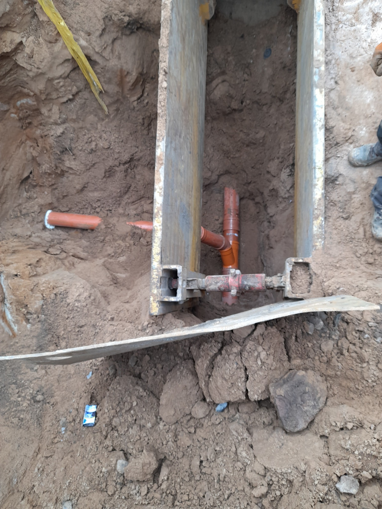 Pomarańczowe rury kanalizacyjne w wykopie zabezpieczonym metalowymi szalunkami, widoczny fragment poziomicy i opakowanie po chusteczkach na ziemi.