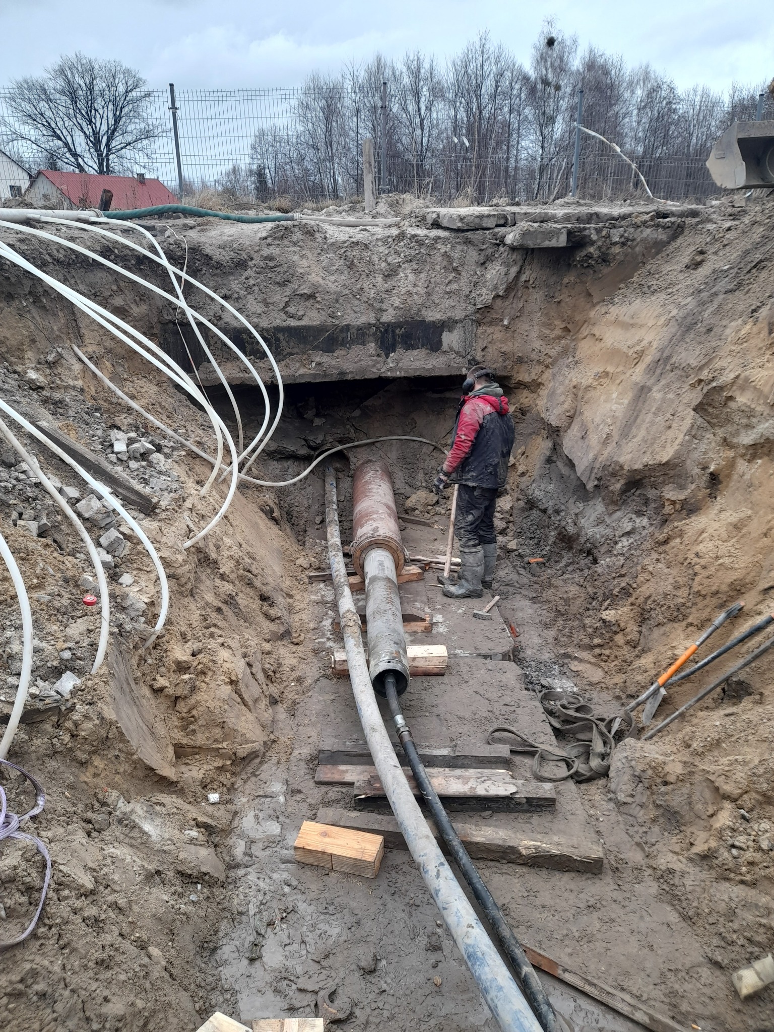 Głęboki wykop w ziemi z ułożonymi rurami i pracownikiem w odzieży roboczej, widoczne narzędzia i białe rury na skraju wykopu.
