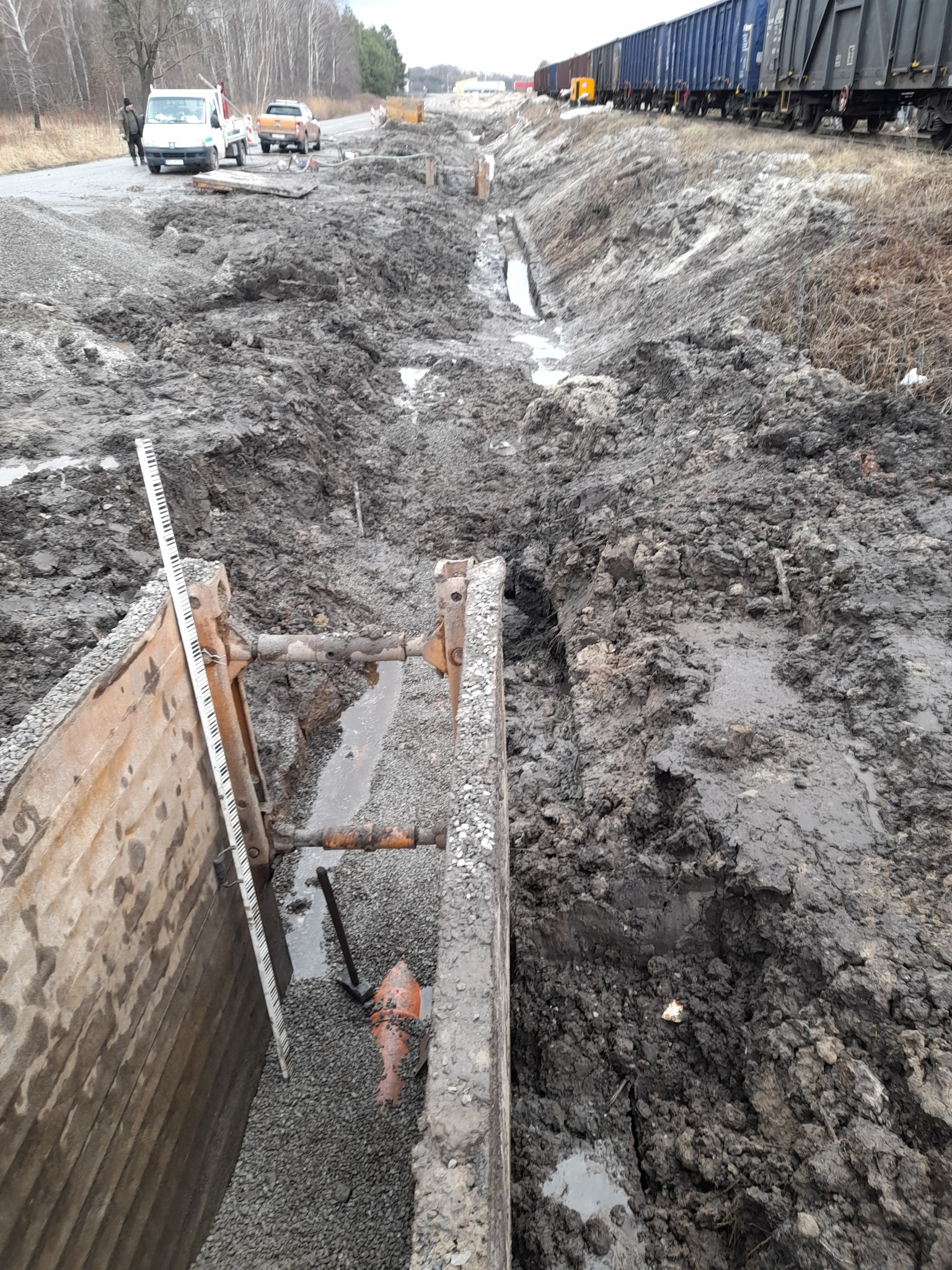 Wykonany wykop z zabezpieczeniem ścianek, widoczna linia kolejowa w tle, teren po deszczu.