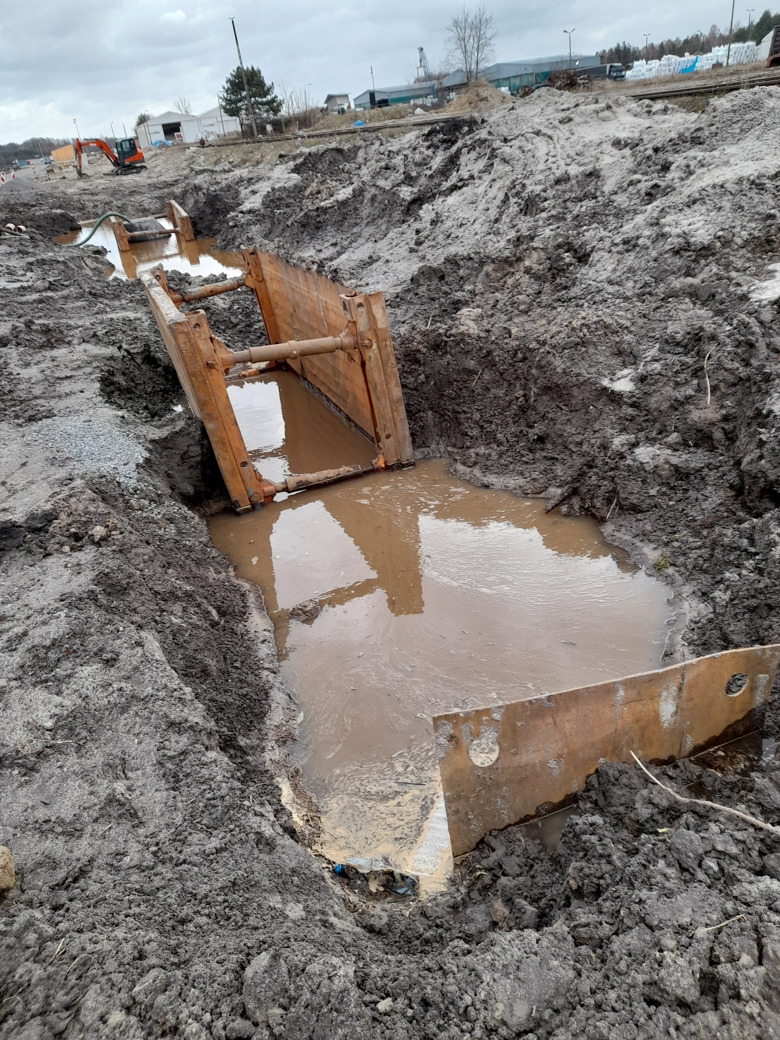 Wykop zabezpieczony metalowymi szalunkami wypełniony wodą, widoczny w oddali pomarańczowy mały koparka, teren budowy.