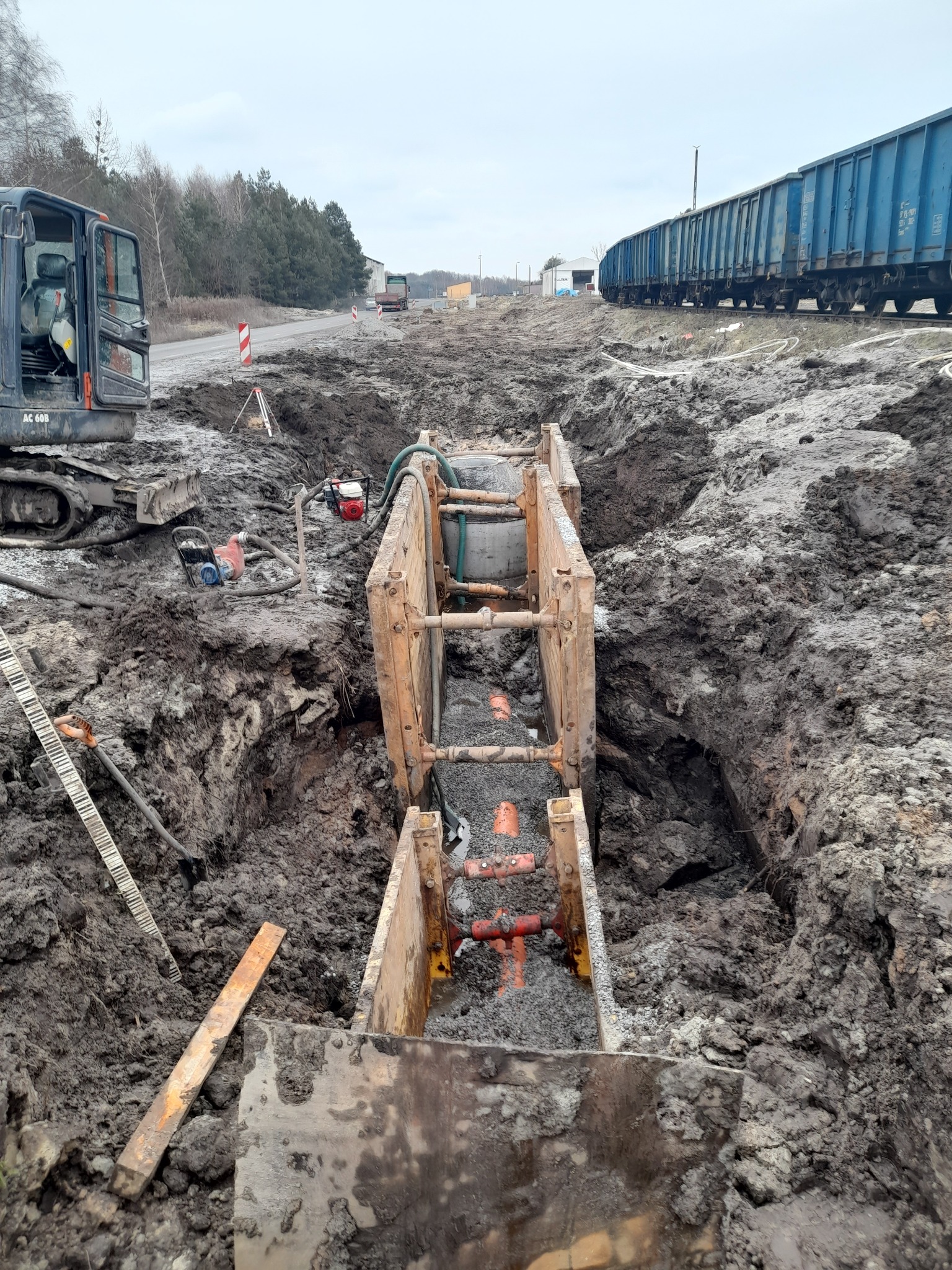 Głęboki wykop zabezpieczony szalunkiem, wewnątrz ułożone rury kanalizacyjne, obok mini koparka, w tle droga i pociąg towarowy.