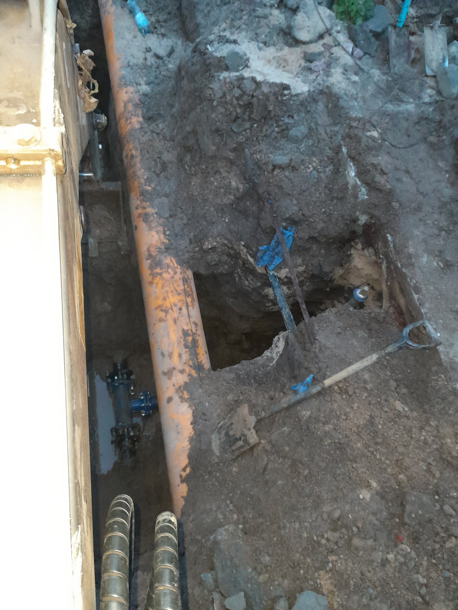 Wykop z widoczną rurą wodociągową z brązowym nalotem, złączkami, leżącą obok łopatą i fragmentem maszyny wykopowej po lewej stronie.