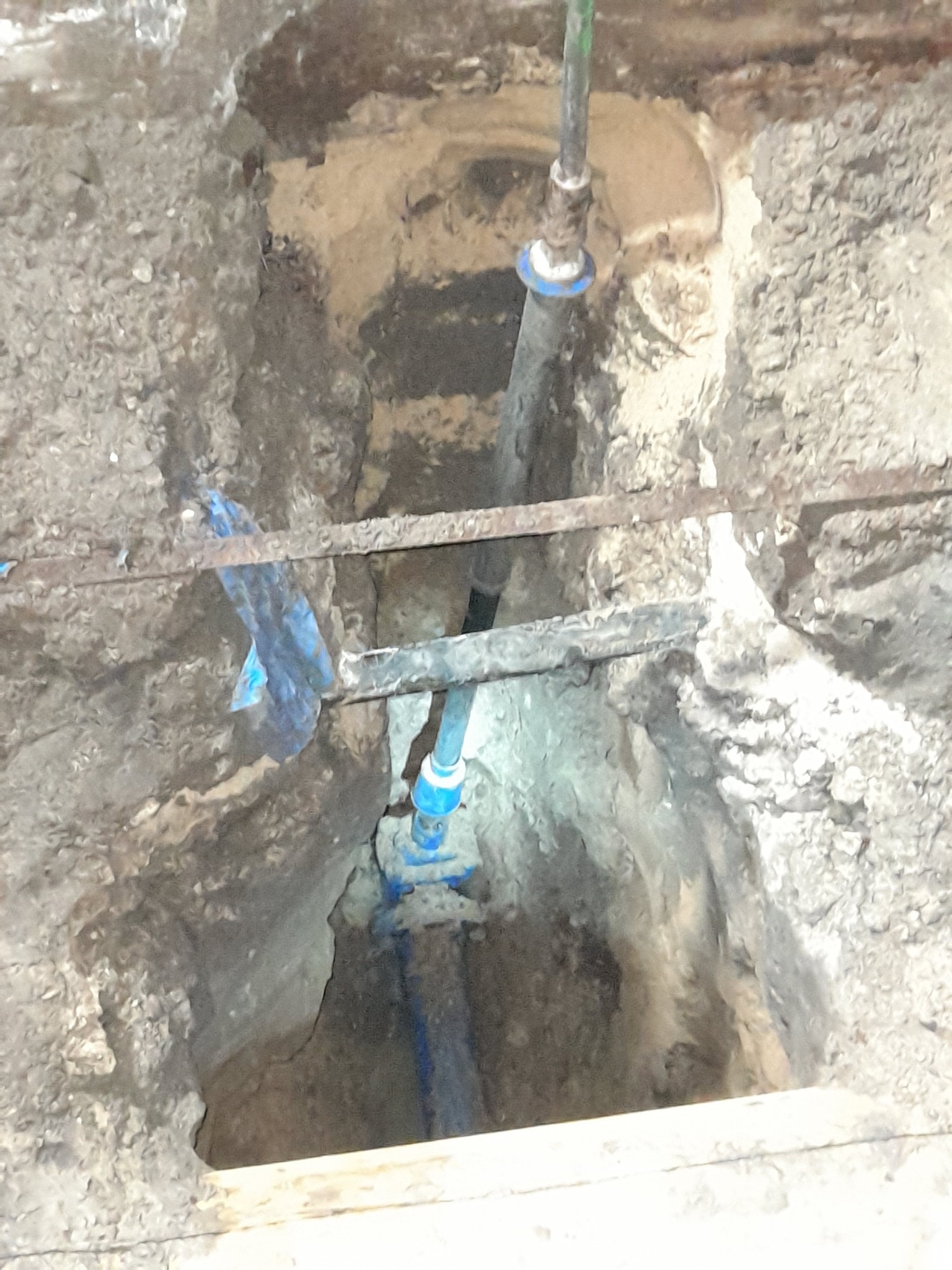 Głęboki wykop w ziemi z widocznymi rurami instalacji wodnej z niebieskimi złączkami, częściowo oświetlony, z metalowymi wzmocnieniami i fragmentem niebieskiej folii.