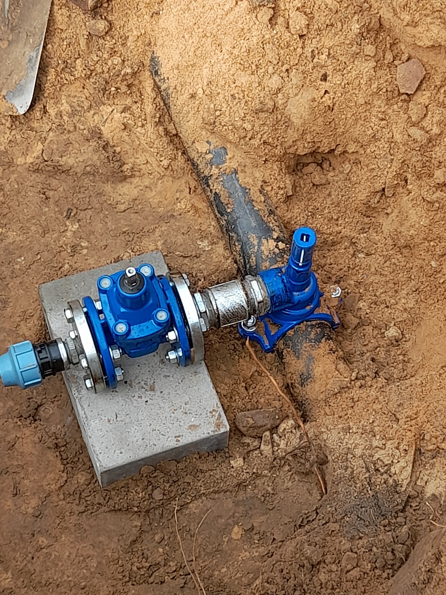 Niebieski zawór z metalowymi połączeniami na betonowym bloczku, podłączony do czarnej rury w wykopie z ziemi.