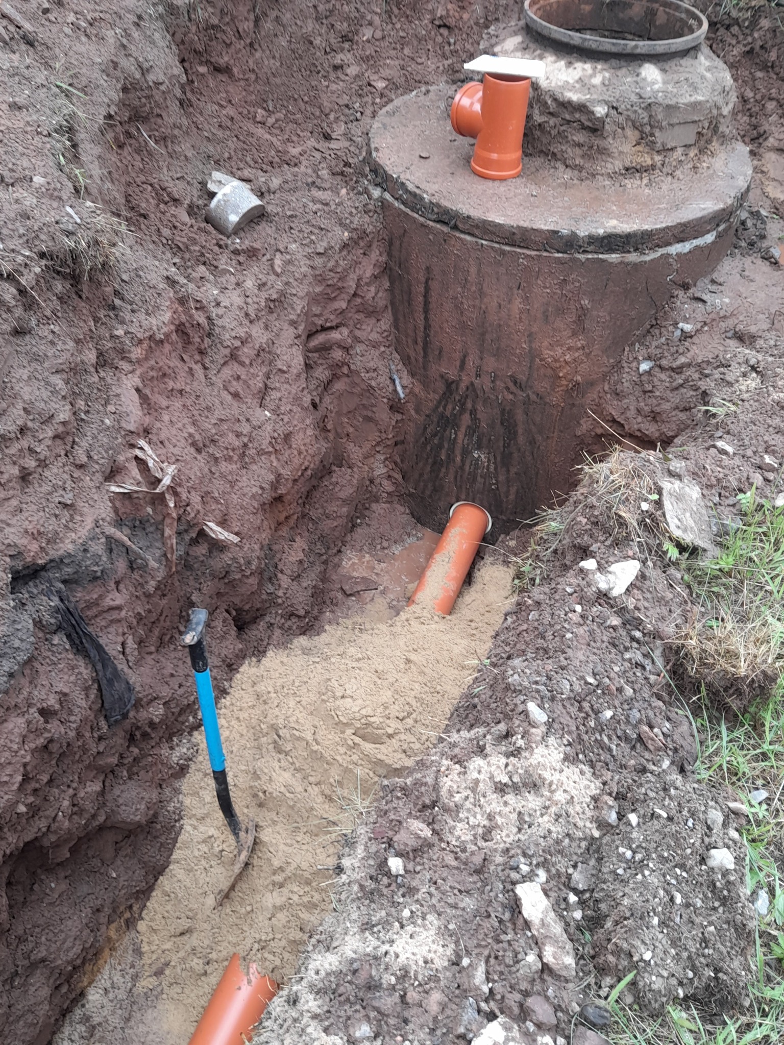 Wykop z betonowym kręgiem, rurami kanalizacyjnymi z tworzywa, zasypany piaskiem, z widoczną łopatą z niebieskim trzonkiem.