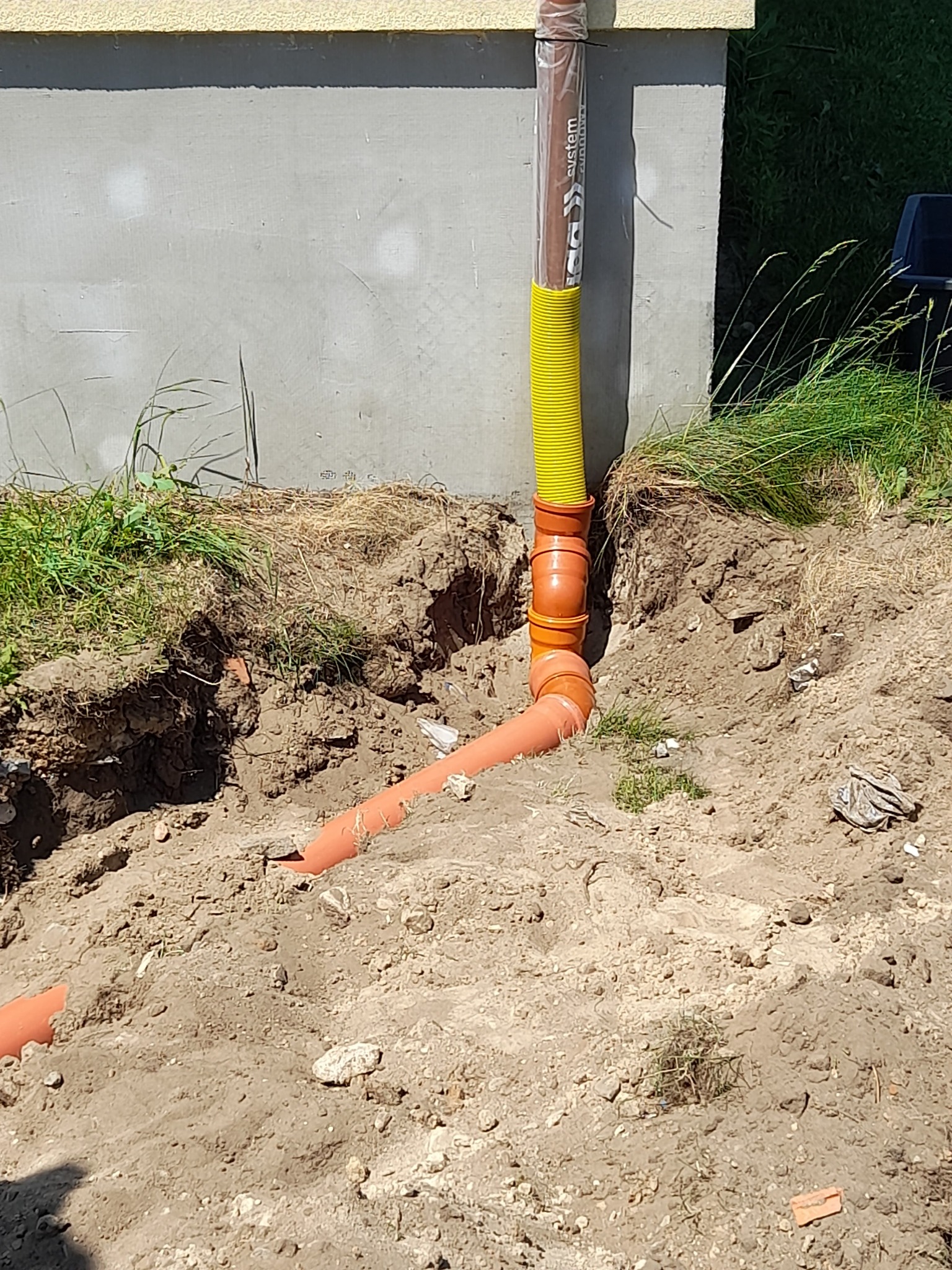 Pomarańczowe rury kanalizacyjne z żółtą osłoną ochronną wystają z ziemi obok fundamentu budynku, widoczne ślady wykopu.