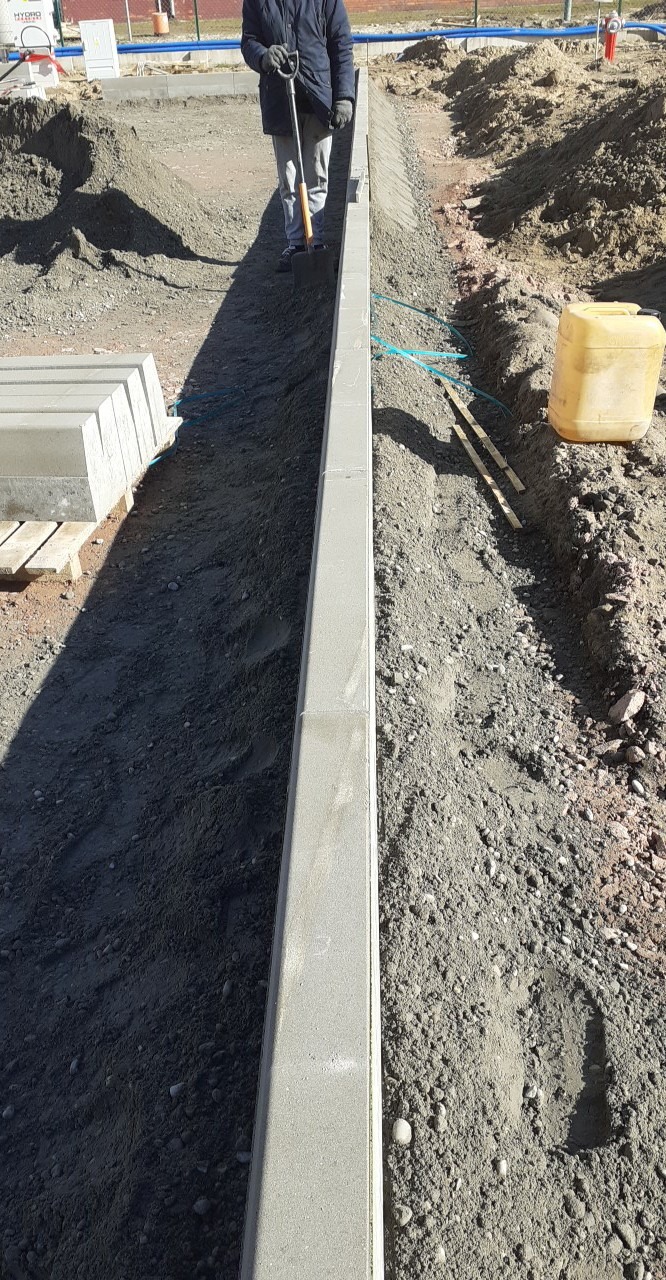 Montaż obrzeży chodnikowych z szarego betonu, widoczny pracownik z łopatą, przygotowane podłoże z piasku i żwiru, paleta z obrzeżami w tle.