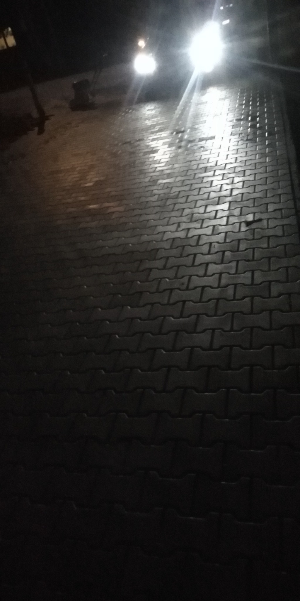 Ułożona kostka brukowa w ciemności, oświetlona przez reflektory pojazdu w oddali, lekko mokra powierzchnia.