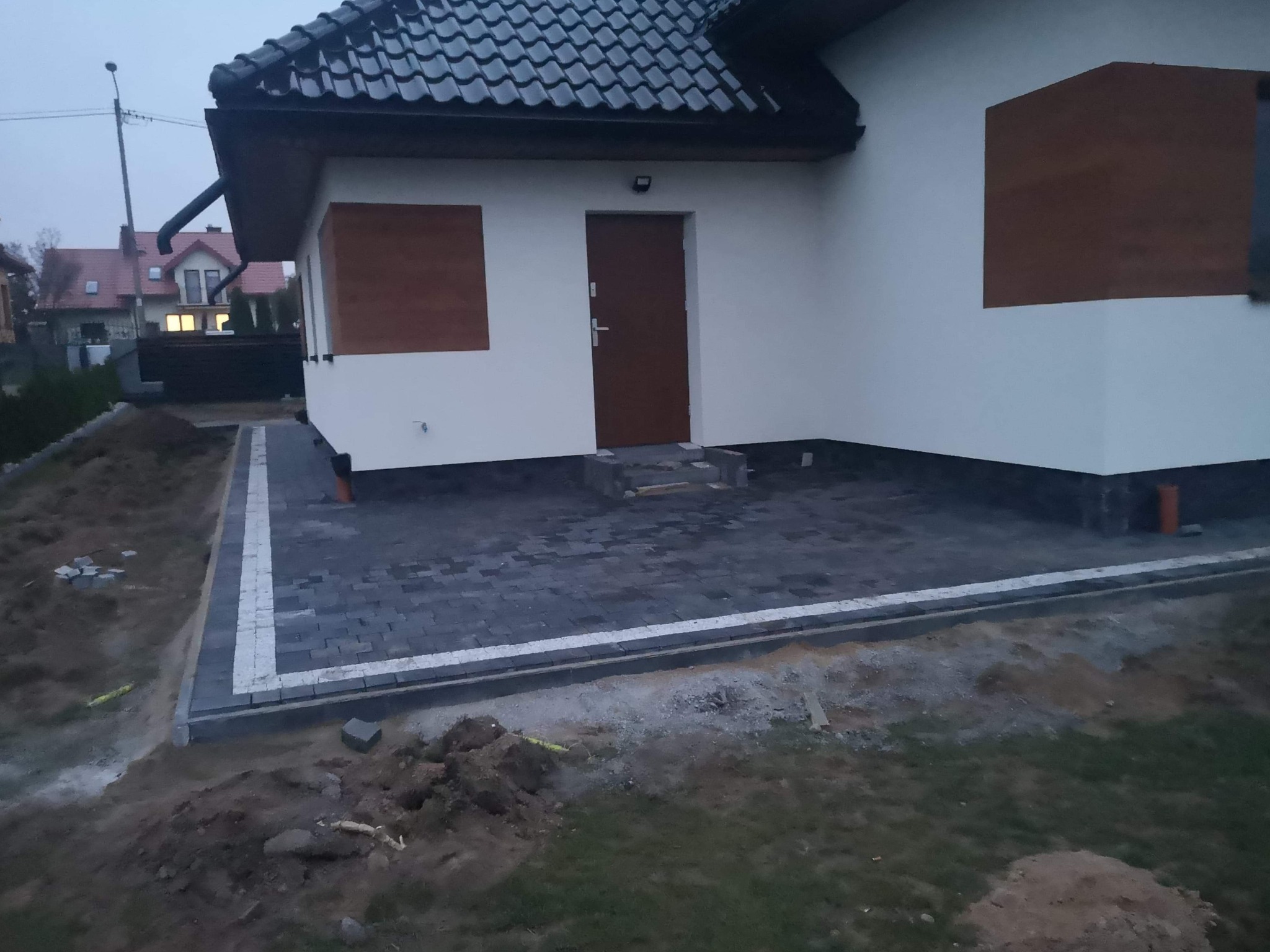 Nowo ułożona, grafitowa kostka brukowa z białym obramowaniem przed domem z brązowymi okiennicami i drzwiami wejściowymi, widoczny fragment trawnika i ziemi przed ułożeniem kostki.