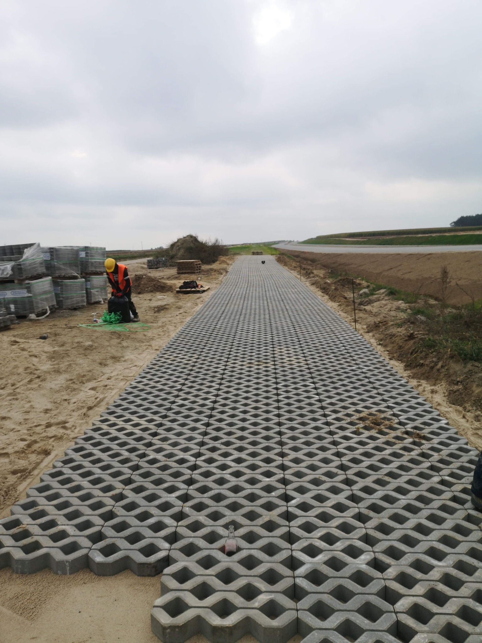 Układanie przepuszczalnej nawierzchni z ażurowych płyt betonowych na piasku, pracownik w kamizelce odblaskowej i kasku przygotowuje materiały, widoczne stosy płyt i krajobraz w tle.