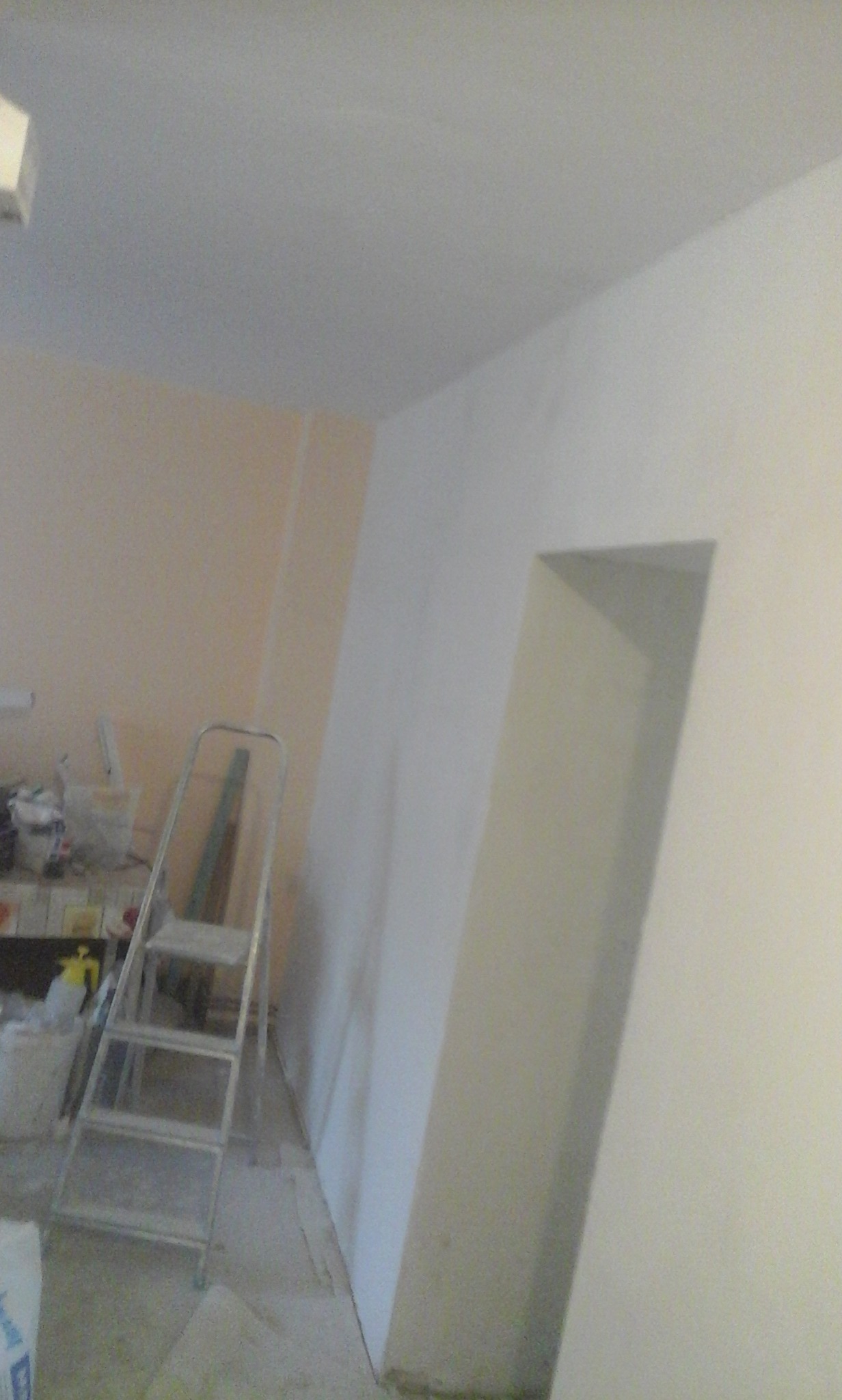 Wnętrze w trakcie prac wykończeniowych: ściana częściowo pomalowana na biało, druga w odcieniach brzoskwini, metalowa drabina oparta o ścianę, widoczne narzędzia i materiały malarskie na stole...