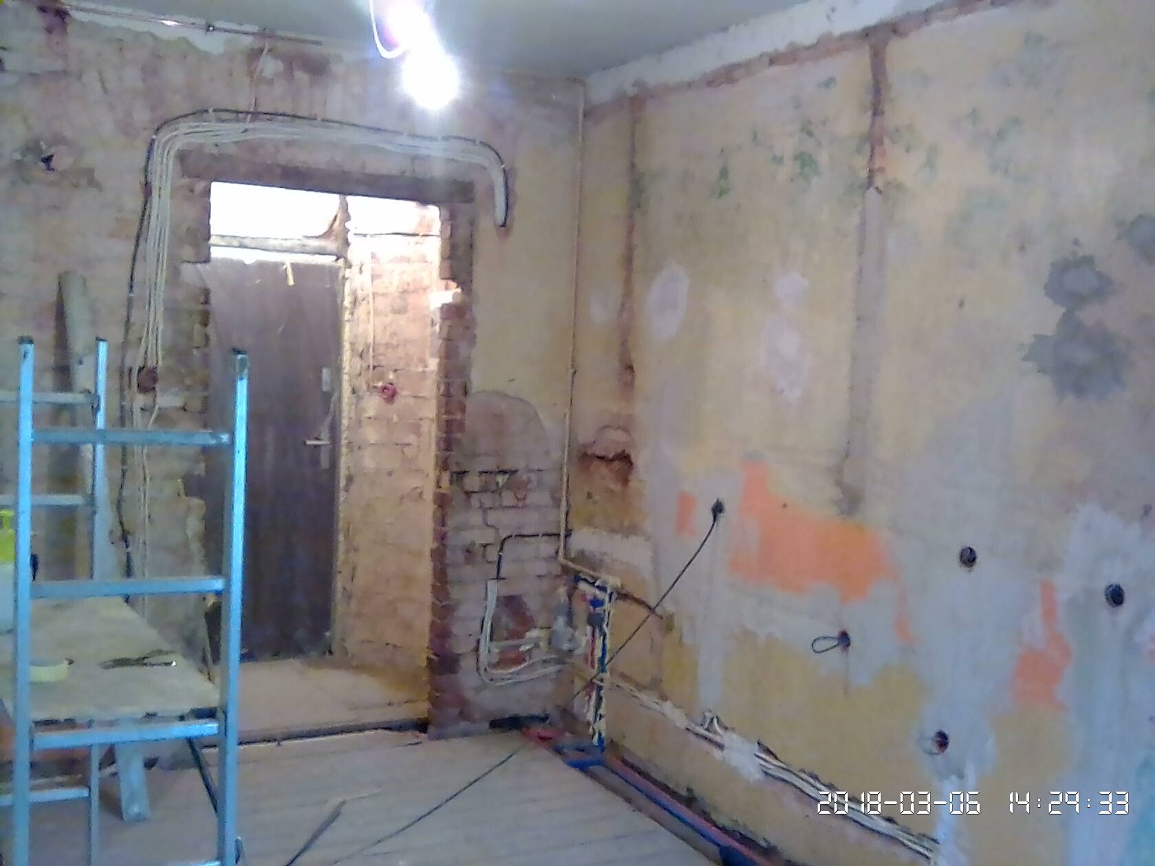 Wnętrze w trakcie remontu: odsłonięta cegła przy ościeżnicy drzwiowej, widoczne przewody elektryczne, rury instalacyjne, fragment ściany z pozostałościami farby i otwory po gniazdkach, drabina...