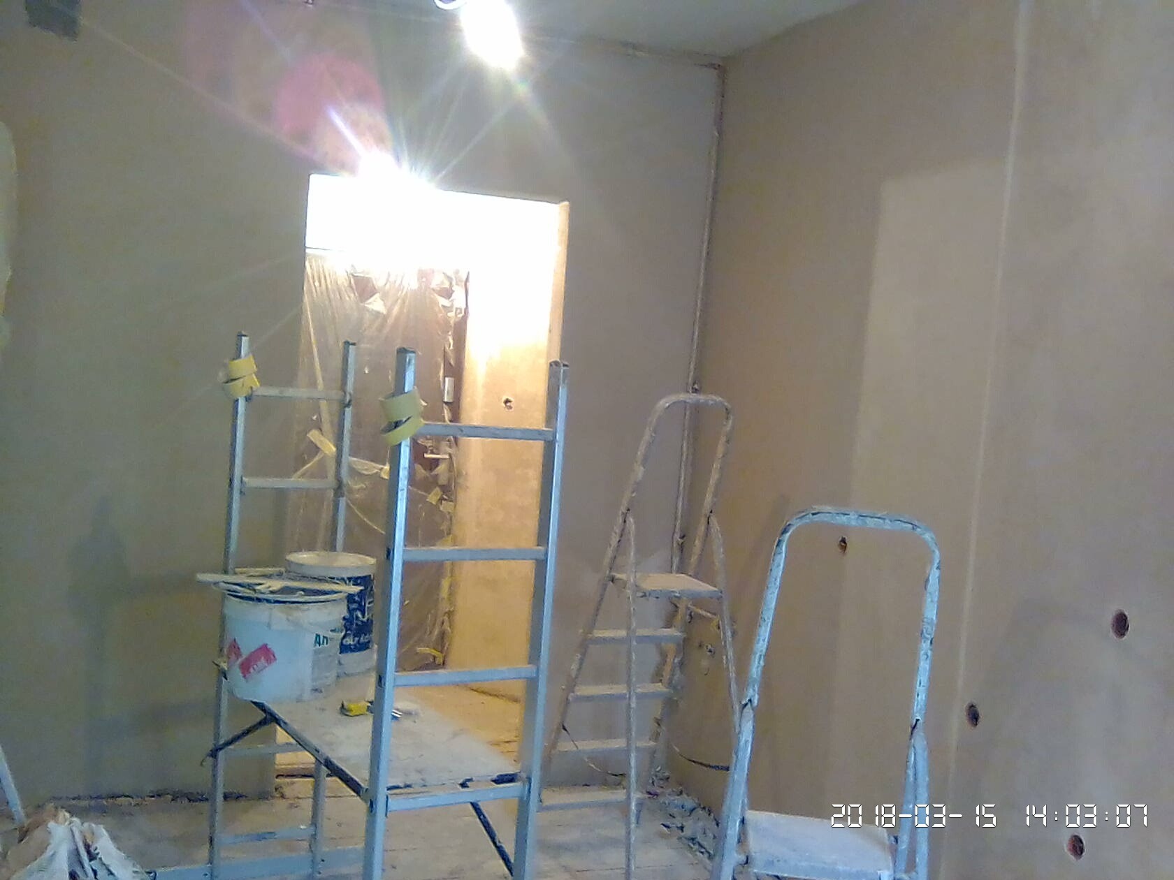 Pokój w trakcie szpachlowania: ściany pokryte świeżą warstwą szpachli, widoczne drabiny malarskie i wiadro z farbą na podeście.