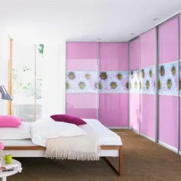 Różowa szafa przesuwna z motywem kwiatowym w sypialni, widok z boku łóżka z lampką nocną i kwiatami w wazonie.