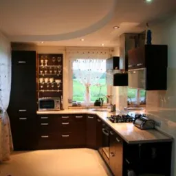 Nowoczesna kuchnia w odcieniach brązu i beżu, z błyszczącą podłogą, oświetleniem punktowym i widokiem na zieleń za oknem. Na blacie kuchennym widoczny czajnik i płyta gazowa.