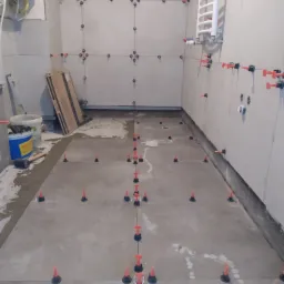 Łazienka w trakcie układania szarych płytek wielkoformatowych na ścianach i podłodze, z użyciem systemu poziomowania płytek (czerwone kliny i czarne podstawki). Na ścianie widoczny grzejnik...