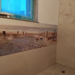 Ściana łazienki wyłożona płytkami z motywem plaży i morza; widoczne przyłącza wodne, okno z folią ochronną i fragment płytek imitujących marmur.