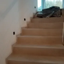 Kamienne schody wewnętrzne w trakcie montażu, zabezpieczone folią ochronną na górnym biegu, z widocznymi puszkami elektrycznymi pod oświetlenie na ścianie.