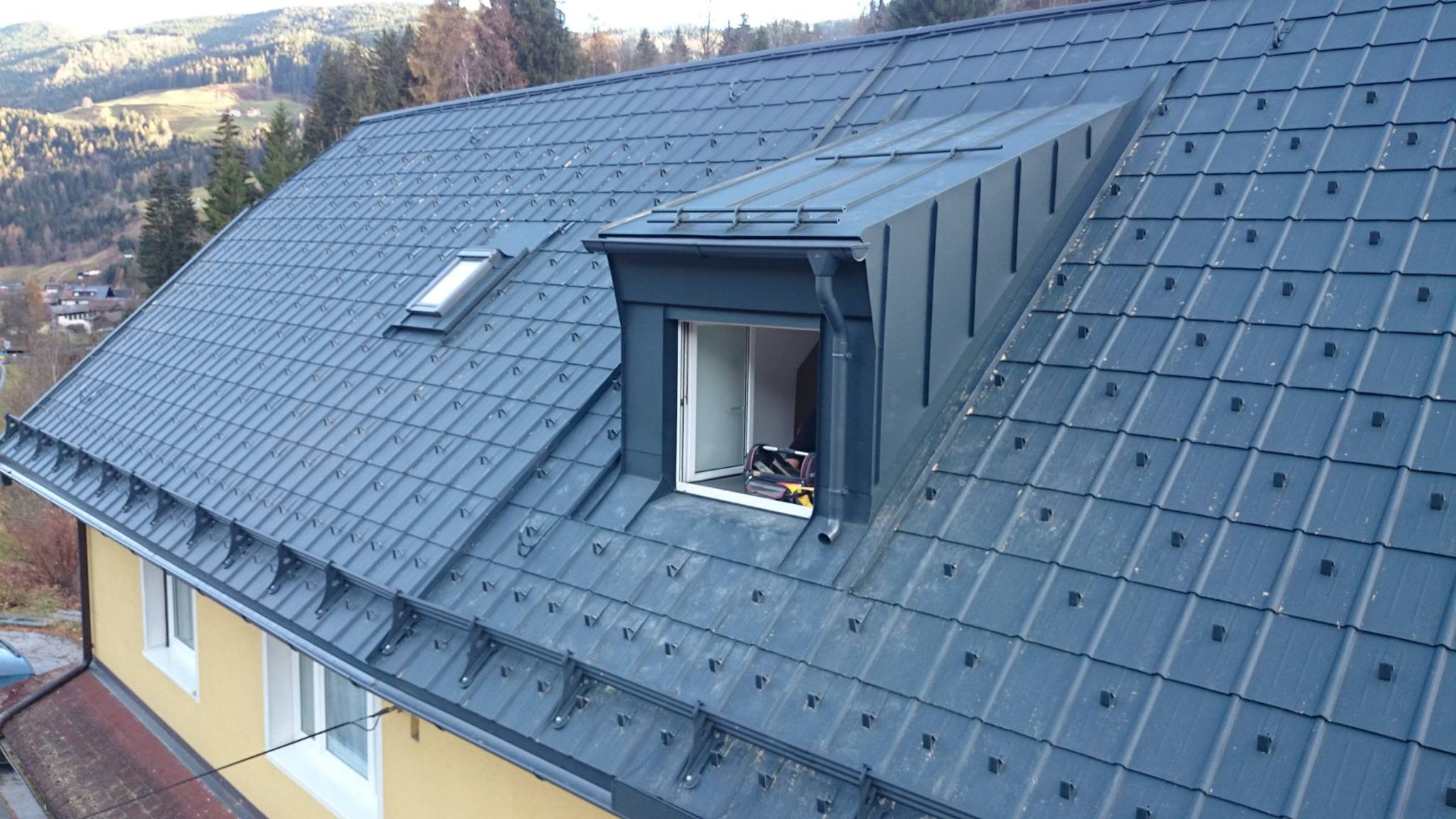 Szary dach z blachy modułowej z oknem dachowym i lukarną, widoczne mocowania blachy, w tle zalesione wzgórza.